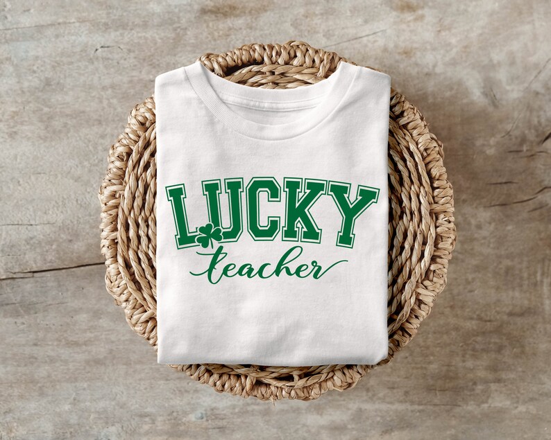 K&ouml;nnte beinhalten: Wei&szlig;es T-Shirt mit gr&uuml;nem Text, der "LUCKY teacher" lautet, mit einem Kleeblatt-Motiv.