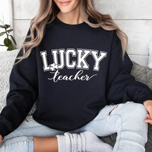 K&ouml;nnte beinhalten: Schwarzer Pullover mit wei&szlig;em Text "LUCKY teacher" und einem Kleeblatt-Motiv.