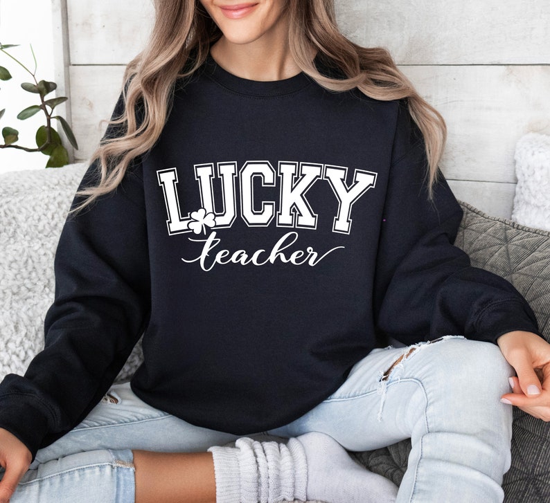 K&ouml;nnte beinhalten: Schwarzer Pullover mit wei&szlig;em Text "LUCKY teacher" und einem Kleeblatt-Motiv.