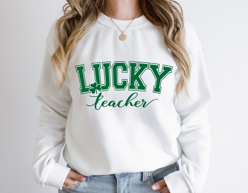 K&ouml;nnte beinhalten: Wei&szlig;er Pullover mit gr&uuml;nem Text, der "LUCKY teacher" mit einem Kleeblatt-Motiv lautet.