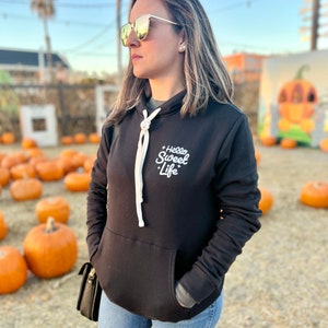 May include: A woman wearing a black hoodie with the text "Hello Sweet Life" in white lettering. She is wearing blue jeans and sunglasses. The background is a field of pumpkins.