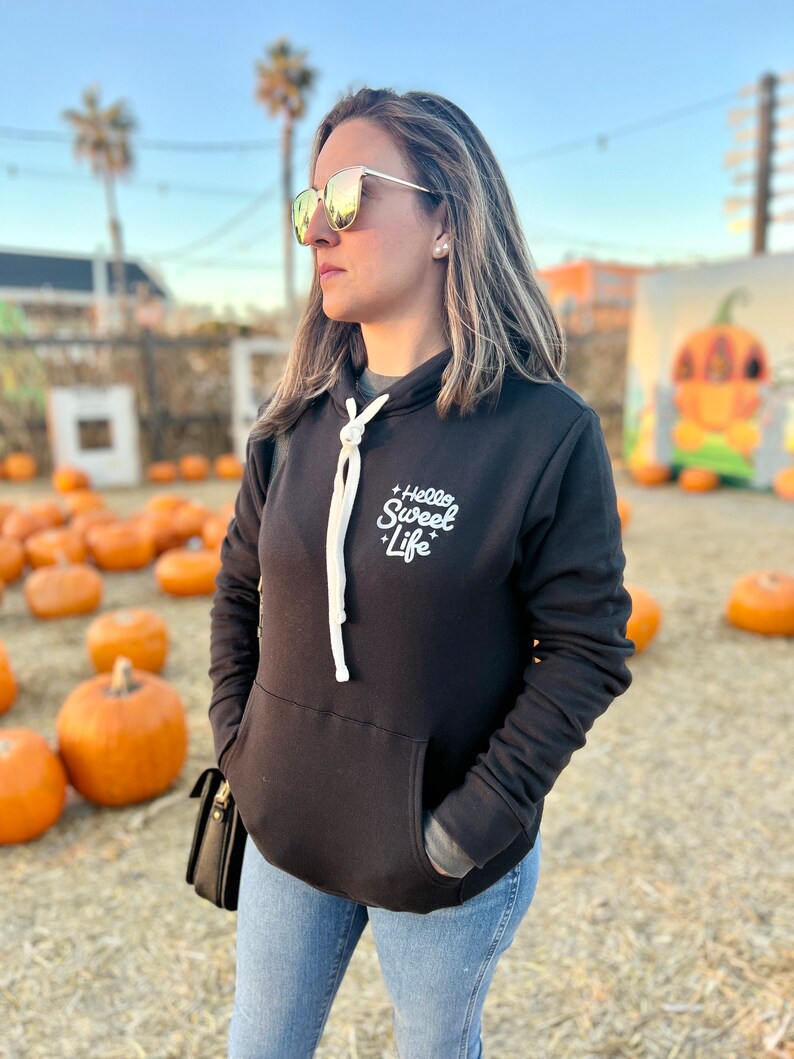 May include: A woman wearing a black hoodie with the text "Hello Sweet Life" in white lettering. She is wearing blue jeans and sunglasses. The background is a field of pumpkins.