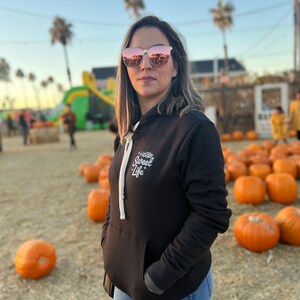 May include: A woman wearing a black hoodie with the text "Hello Sweet Life" in white lettering. She is wearing blue jeans and rose-colored sunglasses. The background is a field of pumpkins.