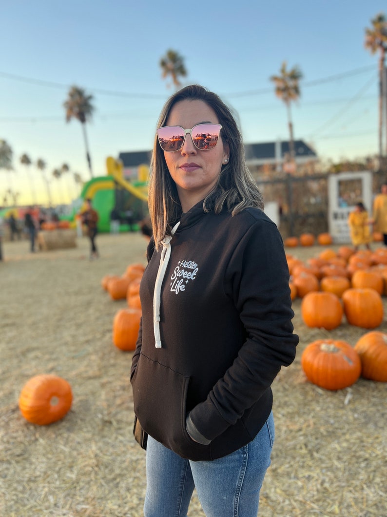 May include: A woman wearing a black hoodie with the text "Hello Sweet Life" in white lettering. She is wearing blue jeans and rose-colored sunglasses. The background is a field of pumpkins.