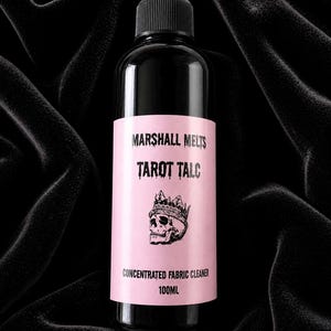Pode incluir: Uma garrafa preta com tampa preta e rótulo rosa. O rótulo diz "MARSHALL MELTS TAROT TALC" com uma caveira usando uma coroa. A parte inferior do rótulo diz "CONCENTRATED FABRIC CLEANER 100ml".