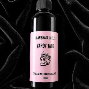 以下が含まれることがあります： 黒いキャップとピンク色のラベルが付いた黒いボトル。ラベルには「MARSHALL MELTS TAROT TALC」と王冠をかぶったスカルが書かれています。ラベルの下部には「CONCENTRATED FABRIC CLEANER 100ml」と書かれています。