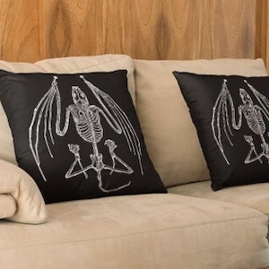 May include: Two black throw pillows with white skeletal bat illustrations. The pillows are on a light beige couch.