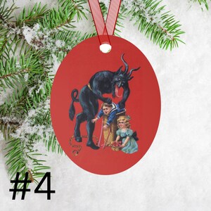 May include: A red Christmas ornament featuring a black devil figure with a long tongue, holding a child in a barrel. A girl in a blue dress stands nearby. The ornament is decorated with a red ribbon.