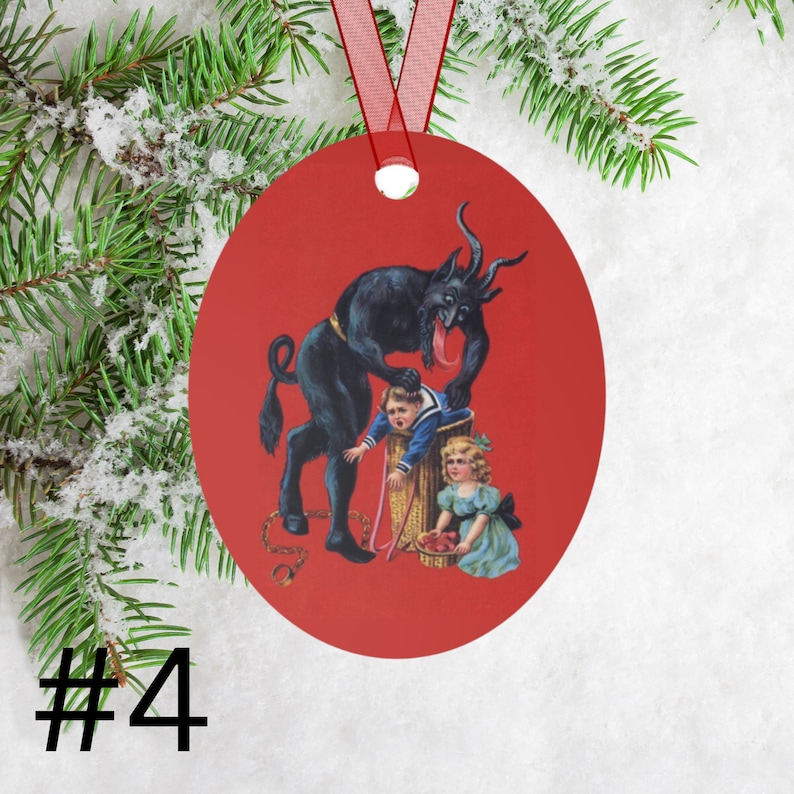 May include: A red Christmas ornament featuring a black devil figure with a long tongue, holding a child in a barrel. A girl in a blue dress stands nearby. The ornament is decorated with a red ribbon.