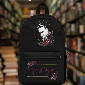 May include: Black backpack with a gold hexagonal frame featuring a black and white portrait of F. Scott Fitzgerald with pink roses. The backpack has a zippered pocket with the signature "F. Scott Fitzgerald" in pink script.