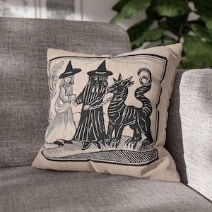 May include: A beige throw pillow with a black and white illustration of two witches and a black beast. The witches are wearing pointed hats and long robes. The beast has a long tail and sharp teeth. The pillow has a black border.
