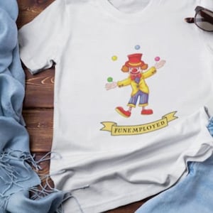 May include: White t-shirt featuring a cartoon clown juggling colorful balls. The clown wears a yellow jacket, red shoes, and a red top hat. The text "FUNEMPLOYED" is on a banner below the clown. Sunglasses are on the top right.