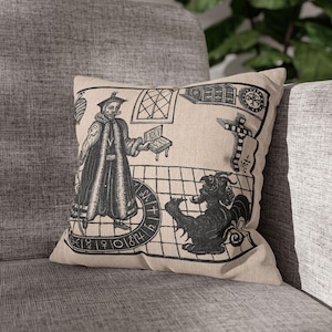 May include: A beige throw pillow with a black and white illustration of a man in a robe standing in front of a circle with numbers and symbols. A black and white illustration of a creature with wings is in the bottom right corner of the pillow.