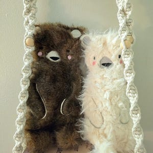 May include: Two plush bear dolls, one brown and one cream, are displayed on a macrame shelf. The bears have embroidered facial features, including closed eyes and pink cheeks. The shelf is suspended by a cream-colored macrame hanger.