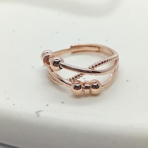 May include: Rose gold adjustable ring with a layered design. The ring features three bands, with beaded and smooth textures, and small ball accents. A stylish piece of jewelry.