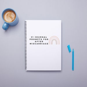 Pode incluir: Um caderno branco de espiral com o texto "31 Journal Prompts for After Miscarriage" e um gráfico de arco-íris. Uma caneca de café azul com café e uma caneta azul estão sobre uma superfície cinza.