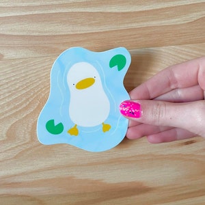 May include: A blue and white sticker with a cartoon duck swimming in a pond. The duck is white with a yellow beak and feet. There are two green lily pads in the pond.
