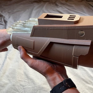 May include: A brown, futuristic arm-mounted device with a clear, spiky projection. The device has a rectangular top with three slots and a circular detail. The design suggests a science fiction or cosplay theme.