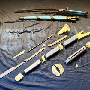 Può includere: Un kit di spada smontato composto da una lama metallica, un fodero blu e bronzo e vari componenti grigi e giallo traslucido. Il set include una guardia circolare, segmenti dell'impugnatura ed estensioni della lama su un tessuto scuro.