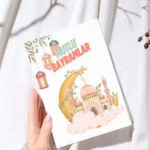 Può includere: Un biglietto bianco con una mezzaluna gialla e una moschea rosa su una nuvola. Il biglietto ha il testo "MUTLU BAYRAMLAR" in lettere verdi e rosa.