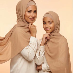 Può includere: Due persone indossano hijab beige e camicie color crema con bottoni. Gli hijab sono drappeggiati sulle loro teste e spalle. Sorridono e guardano la fotocamera. Lo sfondo è di colore pesca chiaro.