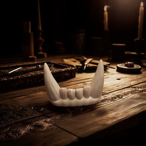May include: A white, sculpted object resembling a set of animal teeth or fangs. The piece features two prominent, elongated pointed fangs on the outer edges, with four smaller, rounded teeth positioned in the center, all connected at the base.