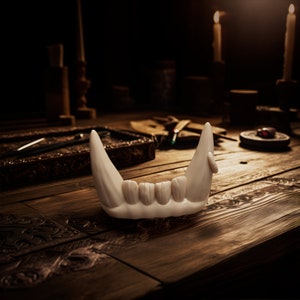 May include: A white, sculpted jawbone piece featuring four small central teeth flanked by two prominent, elongated fangs on either side. The object has a smooth, matte finish and a curved, anatomical shape, presented as a singular decorative item.