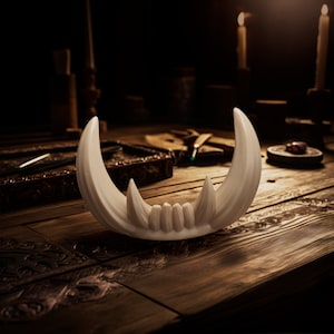 May include: A curved, off-white decorative object shaped like a set of animal tusks or fangs. The piece features two large, pointed outer tusks flanking a central row of smaller, shorter, tooth-like protrusions, all connected at the base.