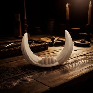May include: A crescent-shaped white object with a smooth, curved surface and a series of small, rounded protrusions along the inner base. The object features two pointed, upward-curving ends that resemble tusks or horns, presented in a solid white color.