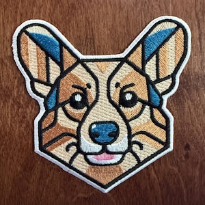 May include: A geometric embroidered patch of a corgi dog with brown, white, and blue colors. The dog is looking directly at the viewer with a friendly expression.
