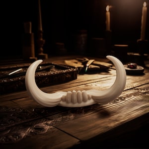 May include: A curved, off-white, tusk-like object featuring a central section with four vertical, rounded ridges. The piece has two symmetrical, pointed ends that sweep upward, creating a crescent shape with a smooth, matte-textured surface.