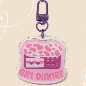 May include: A pink acrylic keychain with a microwave design and the words "Girl Dinner." The microwave has a cow print pattern on top and a heart design on the bottom. The keychain has a silver clasp.