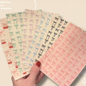 May include: A hand holds a fan of patterned papers. The papers feature repeating patterns of bows and cherries in various colors, including pink, black, blue, and green. Text at the top reads "Colors will vary from images due to differences in device screens."
