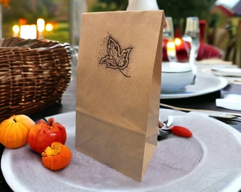 May include: A brown paper bag with a leaf design printed on the front. The bag is sitting on a white plate with a grey design. There are two small orange fruits on the plate.