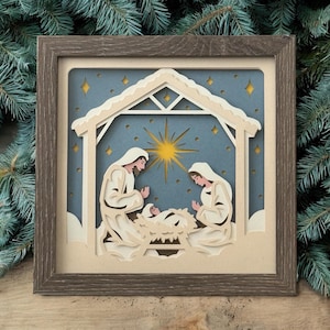 May include: A layered paper art piece depicting the Nativity scene. The artwork features a stable with Mary, Joseph, and baby Jesus, set against a blue starry background. The piece is framed in a brown wooden frame.