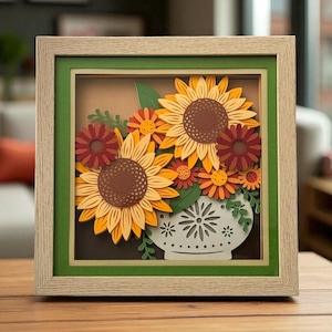 May include: A framed paper art piece featuring a floral arrangement. The artwork showcases sunflowers, daisies, and leaves in shades of yellow, orange, and red, arranged in a white vase with a decorative pattern. The frame is light wood with a green border.
