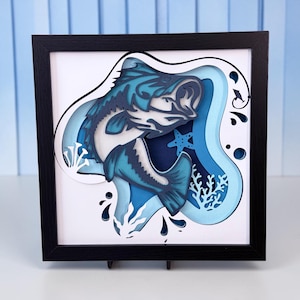May include: A framed 3D paper art piece featuring a leaping blue bass fish. The artwork uses layered paper to create depth, with shades of blue and white. Includes a starfish, coral, and water droplets. The frame is black.