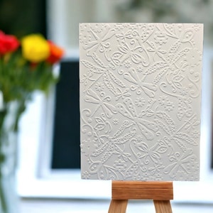 May include: A white, rectangular card with an embossed dragonfly and floral pattern, displayed on a small wooden easel. The card's design features detailed, raised textures. The background includes a blurred view of colorful flowers.