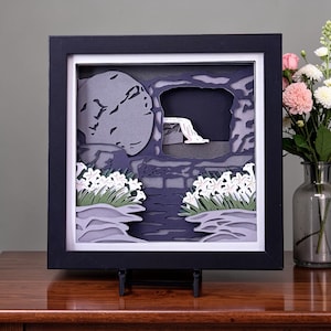 May include: A framed paper art piece depicting a religious scene. The artwork features a dark tomb with a large stone, white lilies, and a dark background. The frame is black, and the artwork is displayed on a stand.