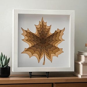 May include: A framed, layered paper art piece featuring a maple leaf design. The leaf is crafted from various shades of brown paper, creating a 3D effect. The artwork is displayed in a white square frame and rests on a black stand.