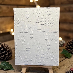 May include: A white embossed card featuring a forest of stylized Christmas trees and snowflakes. The card is displayed on a small wooden easel, with pine cones and holly in the background. The design is a festive, winter-themed craft.