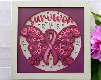 Może przedstawiać: Oprawione, warstwowe dzieło sztuki papierowej o tematyce świadomości raka piersi. Projekt przedstawia różową wstążkę tworzącą skrzydła motyla, ze słowem "Survivor" w skrypcie. Grafika jest w odcieniach różu i fioletu, na białym i fioletowym tle.