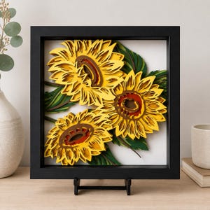 May include: A square black frame containing a multi-layered paper art piece featuring three sunflowers with bright yellow petals, dark brown centers, and deep green leaves. The layered design creates a three-dimensional effect against a white background.