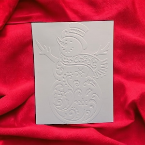 May include: A rectangular white card featuring an embossed snowman design. The snowman is decorated with intricate swirling patterns, snowflakes, and holly leaves. It wears a top hat and a scarf, with its arms extended outward in a cheerful pose.