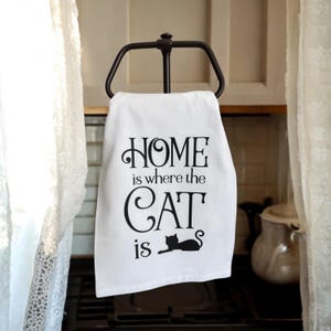 May include: White kitchen towel with black text that reads "Home is where the cat is". A black silhouette of a cat is lying down below the text.