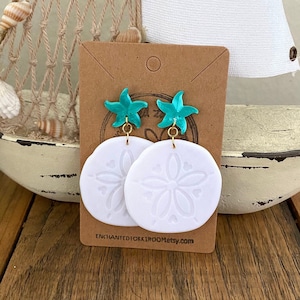 Pode incluir: Brincos de argila branca com um charme de estrela do mar turquesa e um design de d&oacute;lar de areia. Os brincos est&atilde;o em um cart&atilde;o marrom com o texto "ENCHANTED10KK1ROOMetsy.com".