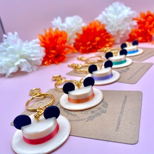Puede incluir: Un conjunto de cinco llaveros con un sombrero blanco con detalles de cinta naranja, morado, azul y verde. Cada sombrero tiene orejas de Mickey Mouse negras y una cadena dorada.