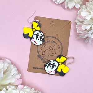 May include: A pair of black and yellow cartoon character earrings with a yellow bow. The earrings are on a brown card with the text "enchanted town" and a rabbit illustration.