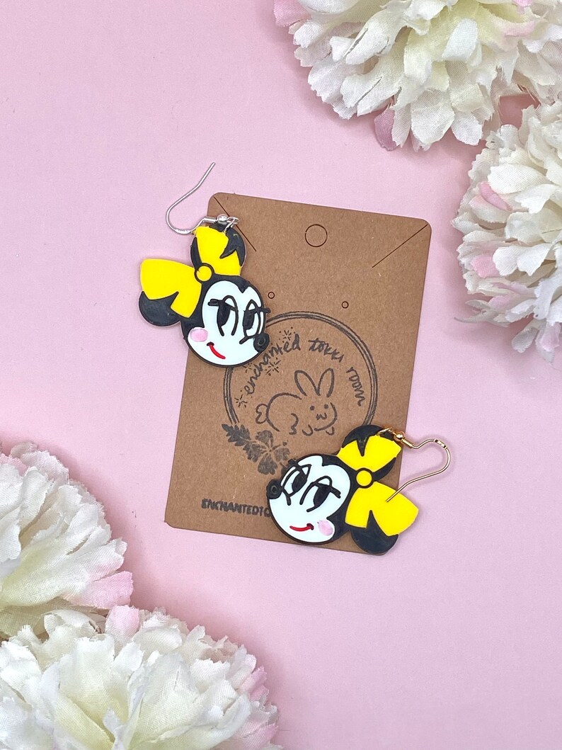 May include: A pair of black and yellow cartoon character earrings with a yellow bow. The earrings are on a brown card with the text "enchanted town" and a rabbit illustration.
