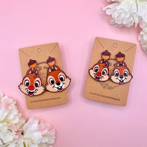 Puede incluir: Un par de pendientes de ardilla marrón con detalles de bellota. Los pendientes están en una tarjeta marrón con el texto "Enchanted Forest Jewelry".